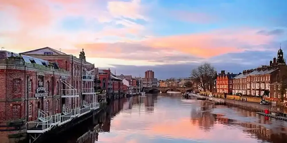 The River Ouse in York at sunset, looking along the water from a bridge. The Bonding Warehouse stands on the left bank, its name lettered on the brickwork above white balconies, with town houses on the right bank and a stone arch bridge in the distance under a pink and blue sky.