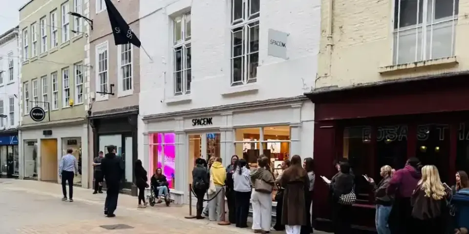 Space NK's new store at St Martins Yard, Coney Street, York, on opening day. Shoppers queue along the pavement beneath the white-painted listed building with its Space NK fascia and projecting sign, with Mango to the left and Rosa's Thai to the right.