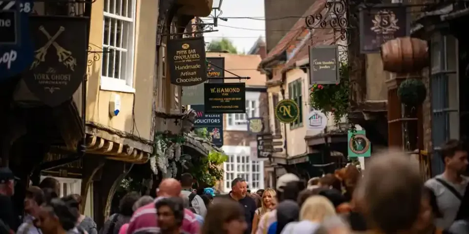 The Shambles, York, on a busy summer day: crowds of shoppers and visitors filling the narrow medieval street, with timber-framed buildings leaning overhead and hanging shop signs for Monk Bar Chocolatiers, W. Hamond and Little Shambles Jewellers.