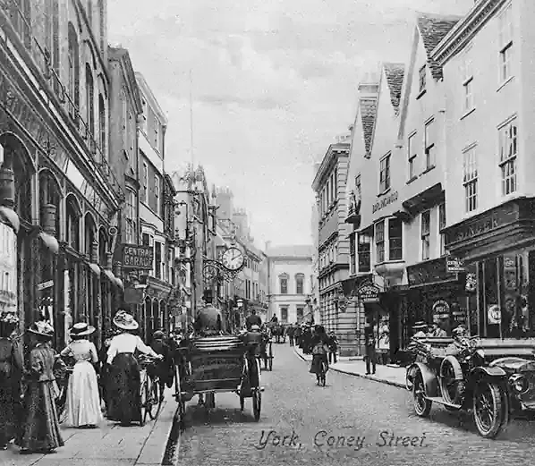 Coney Street, York, in a black and white photograph from the early twentieth century — shoppers in long skirts and wide hats on the pavement, a horse-drawn cart and an early motor car in the cobbled street, and the projecting street clock outside the shopfronts